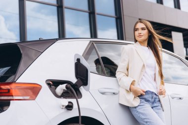 Kendinden emin bir kadın, güneşli bir günde modern ulaşım ve çevre dostu yaşamı simgeleyen şarj istasyonunda elektrikli bir arabaya yaslanıyor..