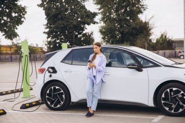 Genç bir kadın akıllı telefonunu kullanırken, şehir ortamında çevre dostu seyahat ve sürdürülebilir enerjiyi kucaklarken, bir şehir istasyonunda elektrikli arabasını şarj ediyor..