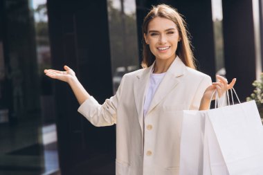 Neşeli bir kadın modern bir perakende mağazasının önünde dururken alışveriş çantalarını neşeyle sergiliyor. Kendine güveni ve mutluluğu şık, açık renkli bir elbiseyle yayar..