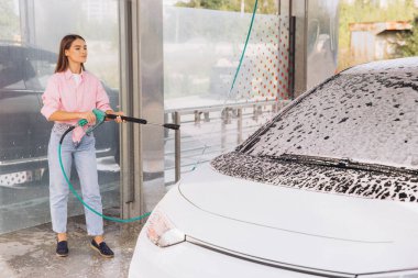 A woman in casual attire uses a pressure washer to clean her car at a self-service car wash. The vehicle is covered in soap, emphasizing cleanliness and maintenance. clipart