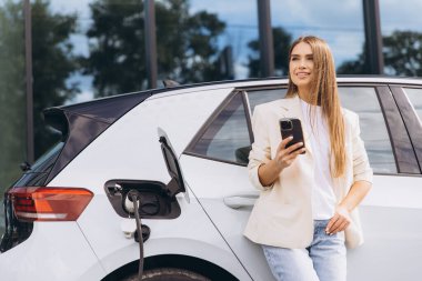 Bir kadın elektrikli arabasının yanında elinde akıllı bir telefonla modern ve sürdürülebilir bir yaşam tarzını sergiliyor. Yüklenen araba, çevre dostu ulaşım ve teknolojide yeniliği temsil ediyor..