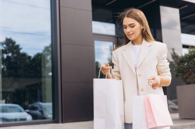 Modern bir binanın dışında yürürken şık bir genç kadın alışveriş torbaları taşır. Şık bir spor ceket giyiyor. Güneşli bir günde moda ve kentsel yaşam tarzını simgeliyor..