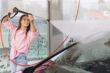 Neşeli genç bir kadın otomobil yıkama istasyonunda elektrikli çamaşır makinesiyle arabasını mutlulukla yıkıyor. Kurulum, bağımsızlık, temizlik ve araç bakımıyla tatmin duygusunu teşvik eder.