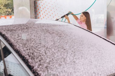 Genç bir kadın oto yıkamada araba temizlemek için köpük sprey kullanıyor. Görüntü araba bakım ve temizlik sürecini yakalıyor.