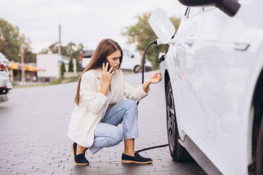 Elektrikli arabanın yanında çömelmiş genç bir kadın telefonuyla konuşurken endişeli görünüyor. O bir açık şehir ortamında. Elektrikli araç sorunları ve yardım kavramı.