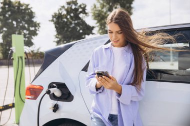 Genç bir kadın şarj olurken elektrikli arabasına yaslanırken akıllı telefonunu kullanıyor. Bu görüntü, modern teknolojiyi, sürdürülebilirliği ve açık hava ortamındaki bağlantıyı yakalar..