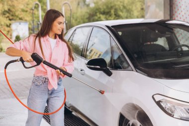 Genç bir kadın açık hava oto yıkama makinesinde arabasını yıkamaktan hoşlanıyor. Gülümsüyor ve günlük giysiler giyiyor, temizlik sırasında neşeli bir atmosfer yaratıyor.