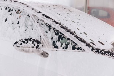 Araba yıkama sırasında kalın sabun köpüğüyle kaplı bir arabanın detaylı görüntüsü. Görüntü temizliği, otomotiv bakımını ve araba yıkama sürecini vurguluyor.