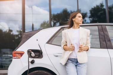 Bir şarj istasyonunda elektrikli bir aracın yanında duran kendine güvenen kadın, modern çevre dostu ulaşımı yansıtıyor. Sahne, kentsel ortamlarda sürdürülebilirlik ve ilerleme hissi yakalıyor.