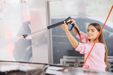 Araba yıkama tesisinde yüksek basınçlı hortumla araba yıkayan gülümseyen genç bir kadın. Sahne, keyifli bir araba bakım rutini sırasında tatmin ve temizlik hissi yakalıyor..