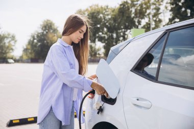 Güneşli bir günde bir otoparkta elektrikli araba şarj eden kadın çevre dostu ulaşım ve sürdürülebilir enerji kullanımı sergiliyor..