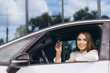 Gülümseyen kadın şık bir arabanın içinde oturuyor, elinde arabanın anahtarlarını tutuyor. Sahne, aydınlık bir günde bir bağımsızlık ve stil hissi yakalıyor..