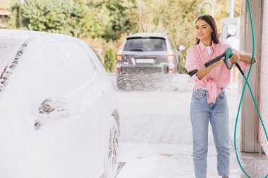 Gündelik giyinmiş genç bir kadın otomatikman araba yıkamada yüksek basınçlı bir hortumla arabasını mutlulukla yıkıyor. Sabunlu su aracı kaplıyor, temizlik sürecini vurguluyor..