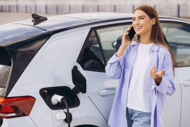 Genç bir kadın elektrikli arabasını şarj ederken ve dışarıda telefonuyla konuşurken gülümsüyor. Görüntü sürdürülebilirlik, teknoloji ve modern yaşam tarzı temalarını yakalar..