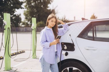 Elektrikli araba şarj istasyonunda akıllı telefonunu kullanan bir kadın. Çevre dostu ulaşım ve modern yaşam tarzını ön plana çıkarıyor. Güneşli bir günde sürdürülebilir yaşamın özünü yakalar.