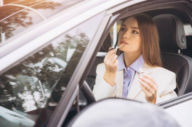 Arabada oturan şık bir kadın, dudak parlatıcısını titizlikle sürüyor. Kendine güveni ve entellektüelliği var. Beyaz ceket ve mavi gömlek giyip bir anlık zarafeti yakalıyor..