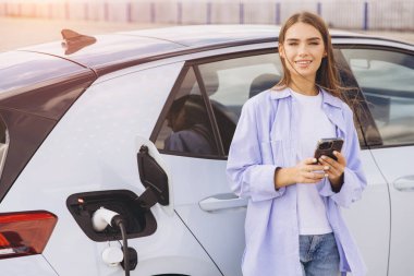 Genç bir kadın elektrikli arabasının yanında gülümsüyor, şehir ortamında akıllı bir telefon tutuyor. Bu görüntü modern teknolojiyi ve çevre dostu yaşam tarzlarını yakalıyor..