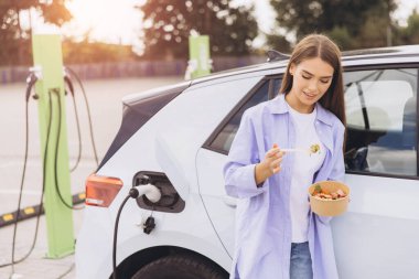 Genç bir kadın, elektrikli arabası istasyonda şarj edilirken sağlıklı bir salatanın tadını çıkarıyor. Öğleden sonra güneş ışığı altında aracın yanında rahatça duruyor..