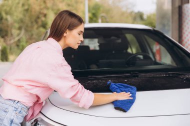 Genç bir kadın araba yıkamada mavi bir bezle arabanın kaputunu siliyor. Pembe bir gömlek ve kot pantolon giyiyor, detaylara ve temizliğe odaklanıyor..