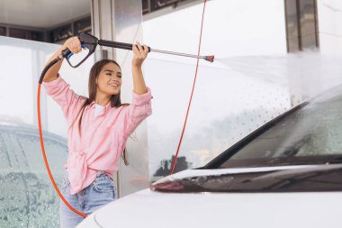 Neşeli genç bir kadın otomobil yıkama istasyonunda elektrikli çamaşır makinesiyle arabasını mutlulukla yıkıyor. Kurulum, bağımsızlık, temizlik ve araç bakımıyla tatmin duygusunu teşvik eder.