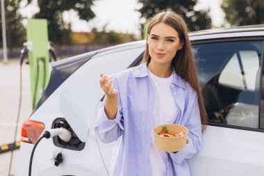 Çağdaş şarj istasyonunda elektrikli arabası şarj olurken genç bir kadın taze salatanın tadını çıkarıyor. Bu resim modern yaşam tarzını ve sürdürülebilir yaşamı yakalar..