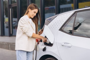 Genç bir kadın açık hava şarj istasyonunda elektrikli arabasını şarj ederken akıllı telefonunu kullanıyor. Modern teknoloji ve çevre dostu ulaşım araçları sergiliyor..