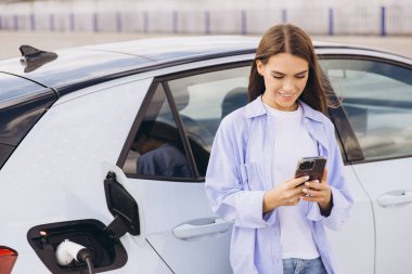 Genç bir kadın elektrikli aracını şarj istasyonunda şarj ederken akıllı telefonunu kontrol ediyor, modern teknoloji ve çevre dostu ulaşım çözümleri sergiliyor..