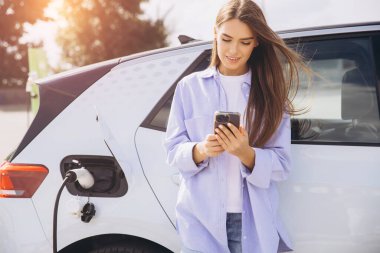 Genç bir kadın elektrikli arabasına yaslanıyor, akıllı telefonunu EV istasyonunda şarj ediyor. Güneş, modern ve sürdürülebilir bir yaşam tarzını yansıtıyor..