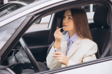 Bir kadın arabasında otururken ruj sürüyor. Açık renkli bir ceket giyiyor ve odaklanmışa benziyor..