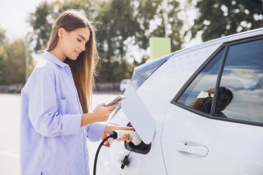 Genç Kadın, güneşli bir günde dışarıda akıllı telefon kullanırken elektrikli arabayı şarj ediyor, Yeşil Enerji ve Sürdürülebilir Yaşam tarzını teşvik ediyor