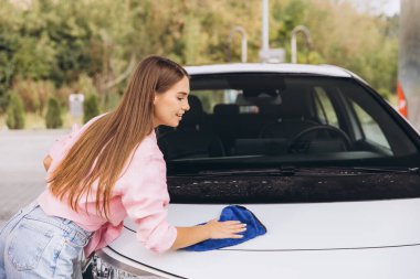 A young woman wipes the car hood with a blue cloth at an outdoor car wash. She wears a pink shirt and jeans, focusing on detail and cleanliness. clipart