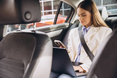 Profesyonel bir kadın, dizüstü bilgisayarına ve telefonuna odaklanarak arabada otururken birden fazla iş yapar. Bu resim, modern uzak çalışma ve dijital bağlantının özünü yakalar..