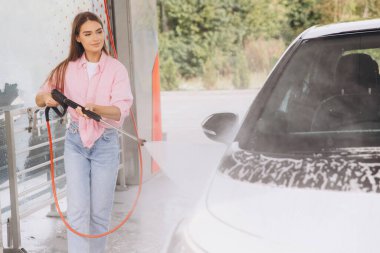 Genç bir kadın oto yıkama servisinde basınçlı yıkama makinesiyle arabasını temizliyor. O, çamaşır yıkama sürecini etkili bir şekilde yönetirken odaklanmış ve memnun görünüyor..