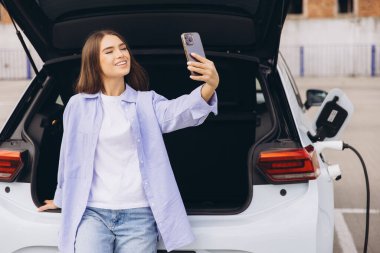 Neşeli genç bir kadın elektrikli arabasının yanında selfie çekiyor, modern bir otoparkta şarj oluyor. Günlük giysiler giyiyor, eko-arkadaşlığı ve çağdaş yaşam tarzını yansıtıyor..