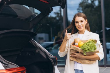 Neşeli genç bir kadın açık bir arabanın bagajında elinde bir torba taze yiyecek ile baş parmak işareti yapıyor. Sağlıklı yaşam tarzı ve alışveriş için ideal..