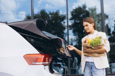 Günlük kıyafetli bir kadın, şehir binasının dışındaki bir cipin bagajından alışveriş yapıyor. Görüntü, market alışverişi ve araba kullanımının rutin bir yaşam tarzını yakalıyor..