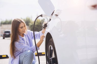 Genç bir kadın titizlikle elektrikli arabasını açık hava şarj istasyonuna bağlıyor. Parlak gün ışığı altında sürdürülebilir enerji çözümlerine olan bağlılığını gösteriyor..