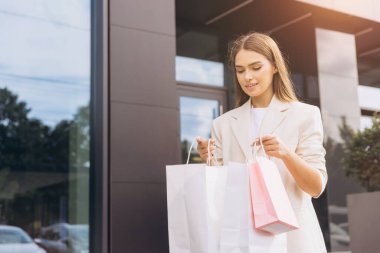 Bej ceketli genç bir kadın dışarıda birkaç alışveriş çantası tutarken gülümsüyor. Sahne, başarılı bir alışveriş yolculuğunun neşe ve memnuniyetini yakalar..