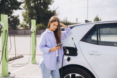Genç bir kadın şarj istasyonunda akıllı telefonunu kullanarak elektrikli arabasına yaslanıyor. Kurulumda modern teknoloji, sürdürülebilirlik ve kolaylık vurgulanıyor.