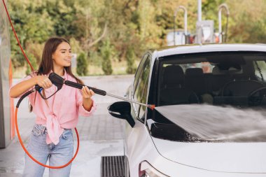 Genç bir kadın açık hava oto yıkama makinesinde arabasını yıkamaktan hoşlanıyor. Gülümsüyor ve günlük giysiler giyiyor, temizlik sırasında neşeli bir atmosfer yaratıyor.
