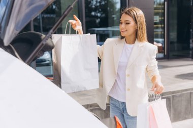 Şık kıyafetli genç bir kadın modern bir binanın dışındaki bagajına mutlu bir şekilde alışveriş torbaları dolduruyor ve başarılı bir alışveriş günü sergiliyor..