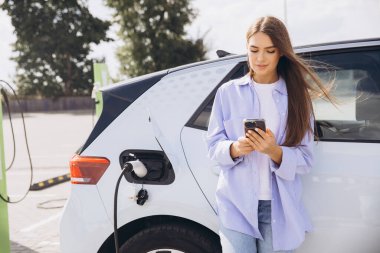 Genç bir kadın şarj olurken elektrikli arabasına yaslanırken akıllı telefonunu kullanıyor. Bu görüntü, modern teknolojiyi, sürdürülebilirliği ve açık hava ortamındaki bağlantıyı yakalar..