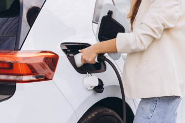 Bir kadın, açık hava istasyonundaki şarj kablosunu fişe takan elektrikli bir arabanın yanında duruyor. Görüntü sürdürülebilir enerji çözümleri ve çevre dostu ulaşım için modern teknolojiyi temsil ediyor.