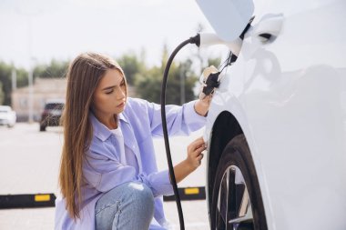 Genç bir kadın titizlikle elektrikli arabasını açık hava şarj istasyonuna bağlıyor. Parlak gün ışığı altında sürdürülebilir enerji çözümlerine olan bağlılığını gösteriyor..
