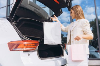 Genç bir kadın alışveriş çantalarını modern bir binanın dışında arabasının bagajına koyar. Şık bir spor ceket giyer ve başarılı bir alışveriş gezisinden zevk alır. Rahatlık ve keyif dolu bir yaşam tarzını yansıtır..