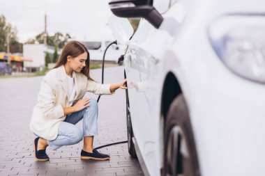 Genç bir kadın açık hava şarj istasyonunda elektrikli arabasını şarj ediyor. Meşgul ve memnun görünüyor, sürdürülebilir enerji ve modern ulaşımı vurguluyor.