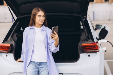 Modern teknolojiyi ve çevre dostu ulaşımı sembolize eden, akıllı bir telefonla elektrikli bir arabaya yaslanmış şık bir genç kadın..