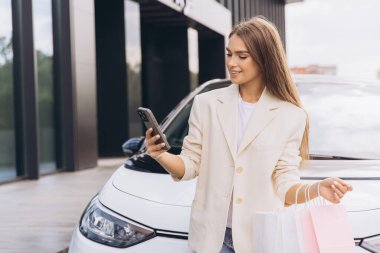 Beyaz bir arabanın yanında dururken akıllı telefonuna gülümseyen alışveriş torbalarıyla genç bir kadın modern yaşam tarzını ve internet bağlantısını temsil ediyor..