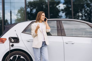 Neşeli bir kadın telefonuyla konuşurken elektrikli arabasının yanında durur. Araba bir şarj istasyonuna bağlı. Sahne modernlik, çevreci dostluk ve bağlılık içeriyor..