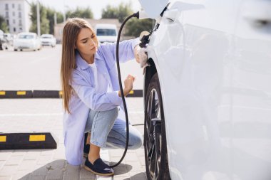 Genç bir kadın elektrikli arabasını şarj istasyonunda şarj ediyor. Bu görüntü çevre dostu teknolojiyi ve güneşli bir günde kentsel bir çevrede sürdürülebilir yaşamı yakalar..